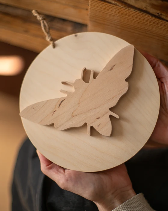 Drewniana dekoracja ścienna w kształcie koła z nałożoną sylwetką ćmy 3D. Rękodzieło wykonane z jasnej sklejki o unikalnym usłojeniu. Produkt zawieszony na naturalnym sznurku, prezentowany w dłoniach na tle drewnianych belek. Niepowtarzalny wzór.