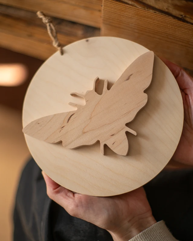 Drewniana dekoracja ścienna w kształcie koła z nałożoną sylwetką ćmy 3D. Rękodzieło wykonane z jasnej sklejki o unikalnym usłojeniu. Produkt zawieszony na naturalnym sznurku, prezentowany w dłoniach na tle drewnianych belek. Niepowtarzalny wzór.