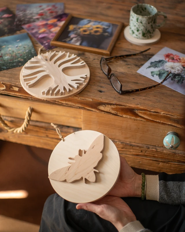 Drewniana dekoracja ścienna w kształcie koła z nałożoną sylwetką ćmy 3D. Rękodzieło wykonane z jasnej sklejki o unikalnym usłojeniu. Produkt zawieszony na naturalnym sznurku, prezentowany w dłoniach na tle drewnianych belek. Niepowtarzalny wzór.